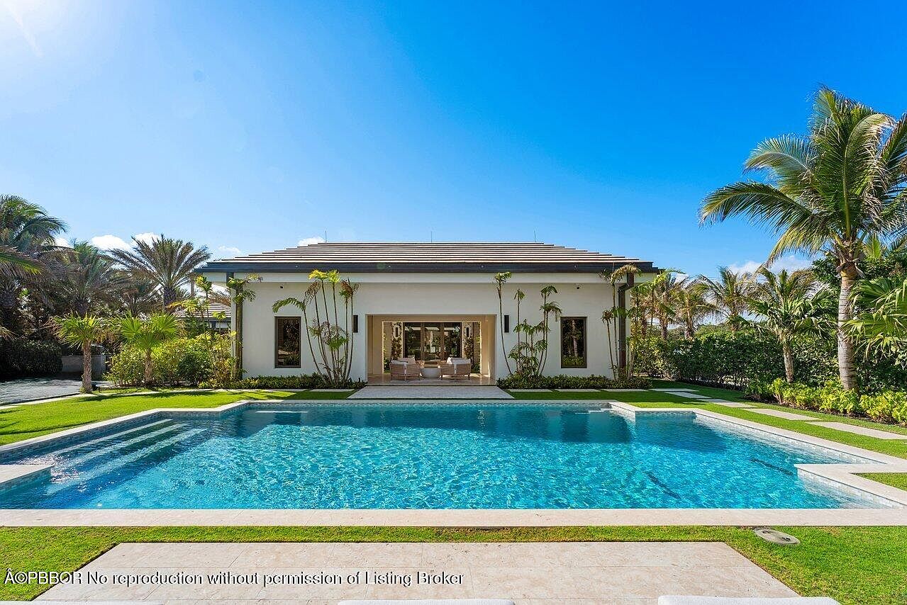 This image showcases a luxurious backyard featuring a large, rectangular swimming pool with clear blue water. The pool is surrounded by well-manicured green grass and lush tropical landscaping, including palm trees. The back of the house is visible, featuring a modern design with large windows and an open patio area, creating an inviting outdoor living space.