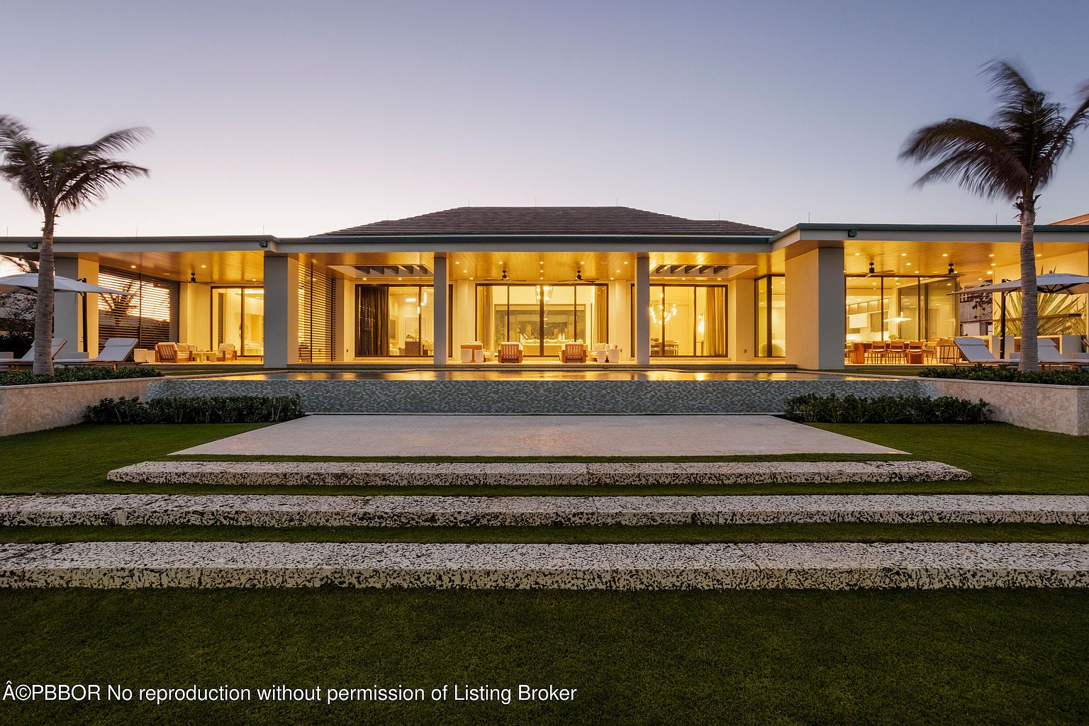 This is a rear exterior view of a luxurious modern home at dusk, featuring a seamless transition between indoor and outdoor living spaces. The property boasts a large pool that reflects the warm interior lighting, a spacious patio area, and meticulously landscaped grounds with palm trees, creating an inviting and serene atmosphere. The architecture emphasizes clean lines and expansive glass doors, enhancing the property's appeal as a high-end real estate offering.