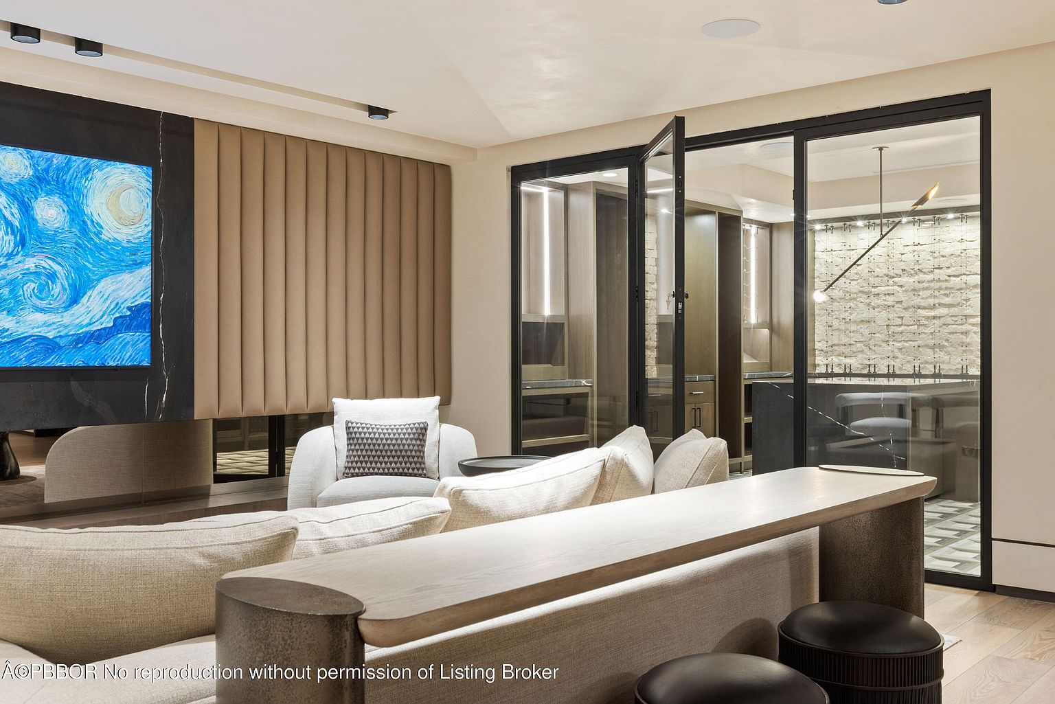 This interior shot showcases a modern living room with a sophisticated design. The room features a large television with a digital art display, a textured wall panel, and comfortable seating arrangements. A unique console table with cylindrical supports adds to the room's contemporary aesthetic, while glass doors offer a glimpse into an adjacent space, enhancing the sense of openness.