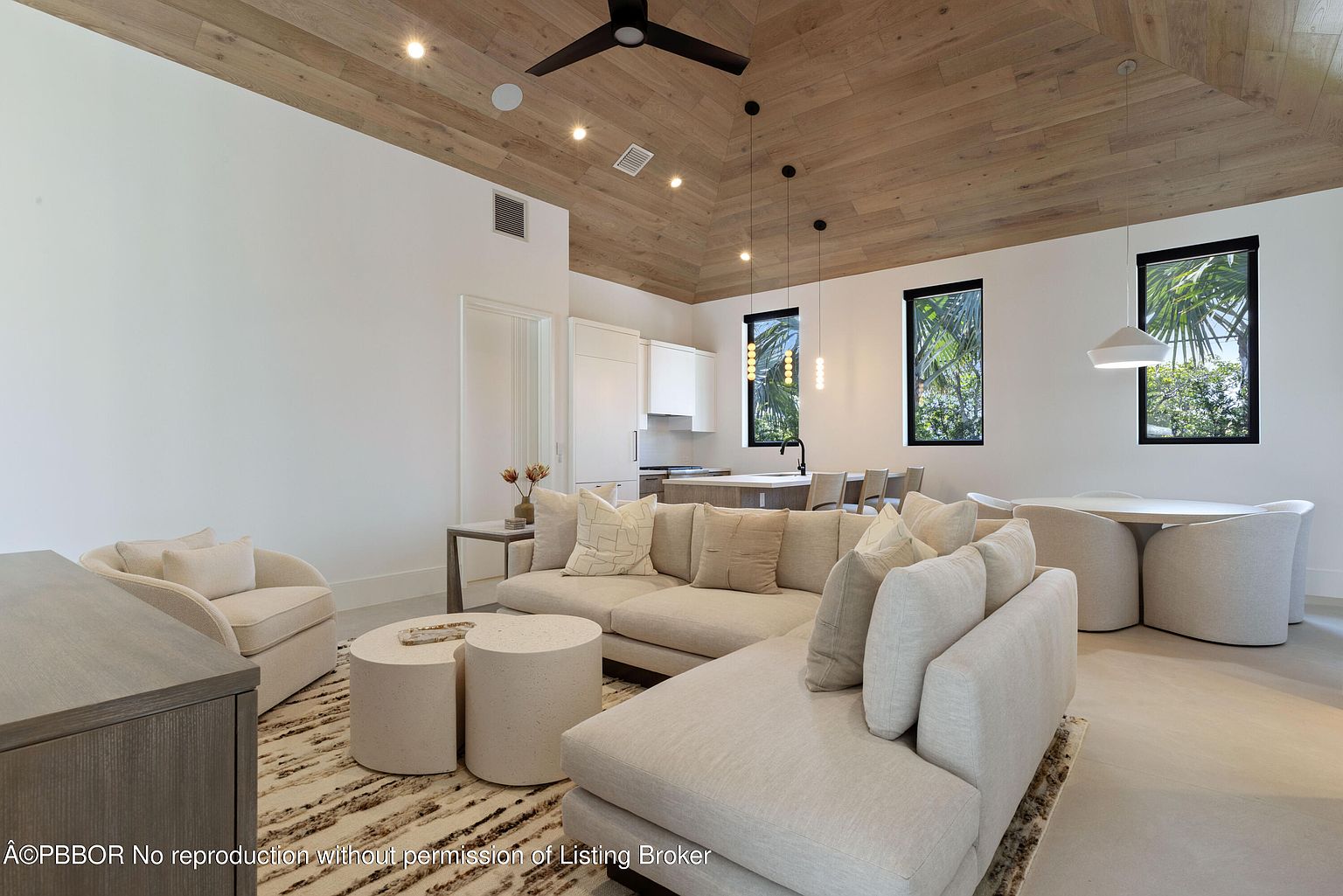 This is an interior shot of a modern living room featuring a large sectional sofa with neutral-toned cushions, complemented by round coffee tables and an accent chair. The room has a light and airy feel, enhanced by the natural wood ceiling, white walls, and natural light streaming through the windows. The open floor plan seamlessly connects the living area to the kitchen and dining space, creating a cohesive and inviting atmosphere.