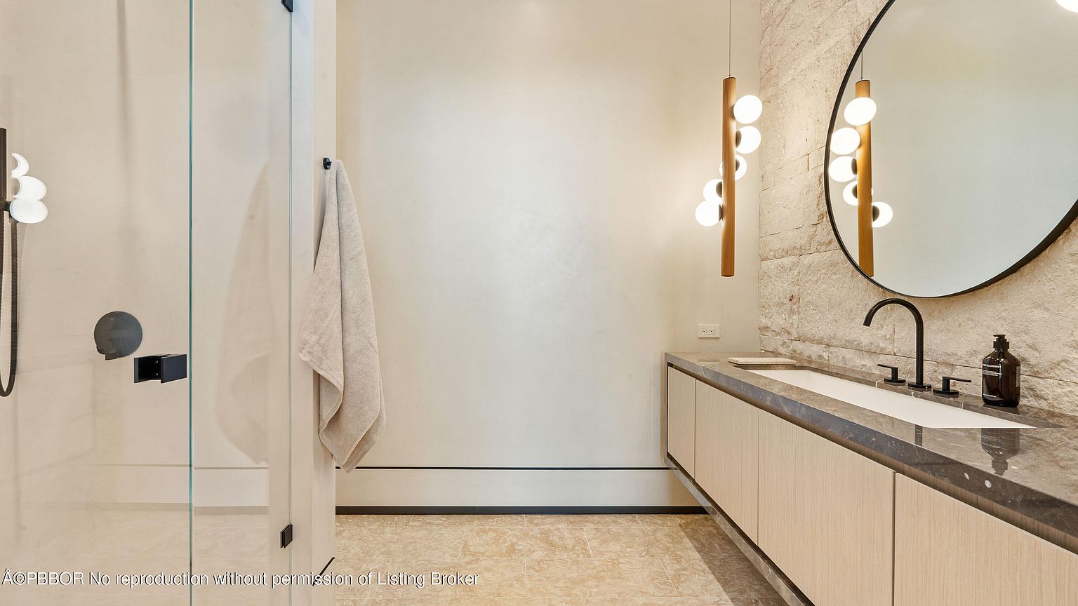 This is a modern primary bathroom featuring a floating vanity with a gray countertop and light wood cabinets. A round mirror hangs above the sink, complemented by a unique pendant light fixture. The bathroom also includes a glass-enclosed shower with matte black hardware, and the walls are a mix of textured stone and smooth plaster, creating a luxurious and contemporary feel.