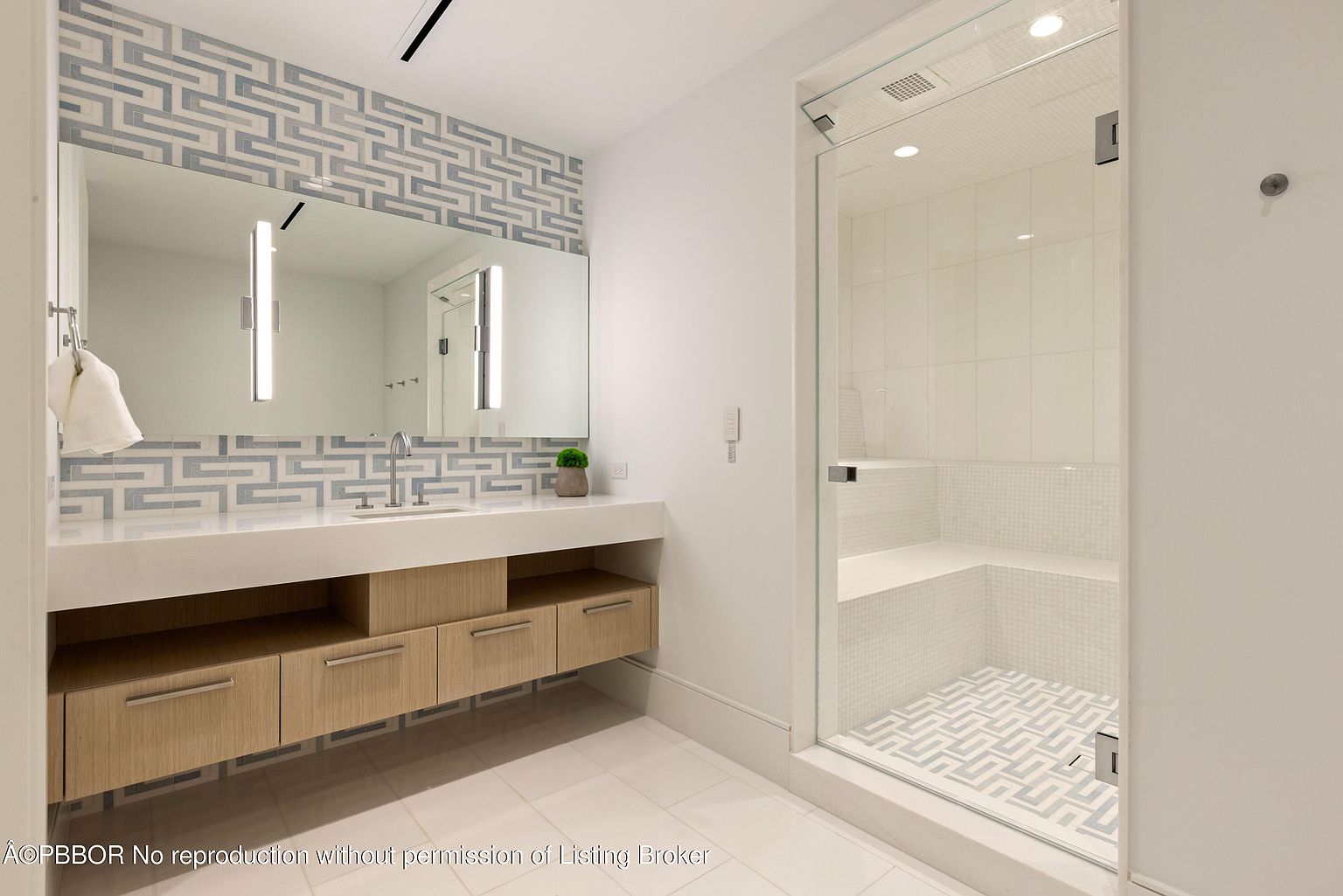 This is a well-lit primary bathroom featuring a modern design with a large mirror above a floating vanity with light wood drawers. The backsplash and accent wall above the mirror showcase a geometric tile pattern, which is echoed in the floor of the glass-enclosed shower. The shower itself has a built-in bench and white tile walls, creating a clean and luxurious space.