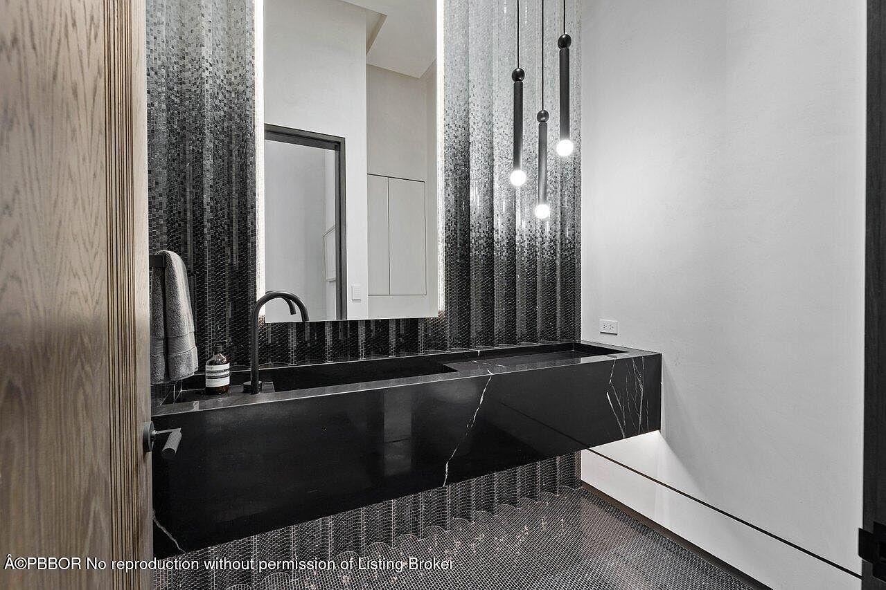 This is a modern bathroom featuring a sleek, black marble countertop with integrated sinks. The wall behind the vanity is adorned with a gradient mosaic tile pattern, complemented by minimalist pendant lighting. The overall design exudes sophistication and contemporary style, creating a luxurious and inviting space.
