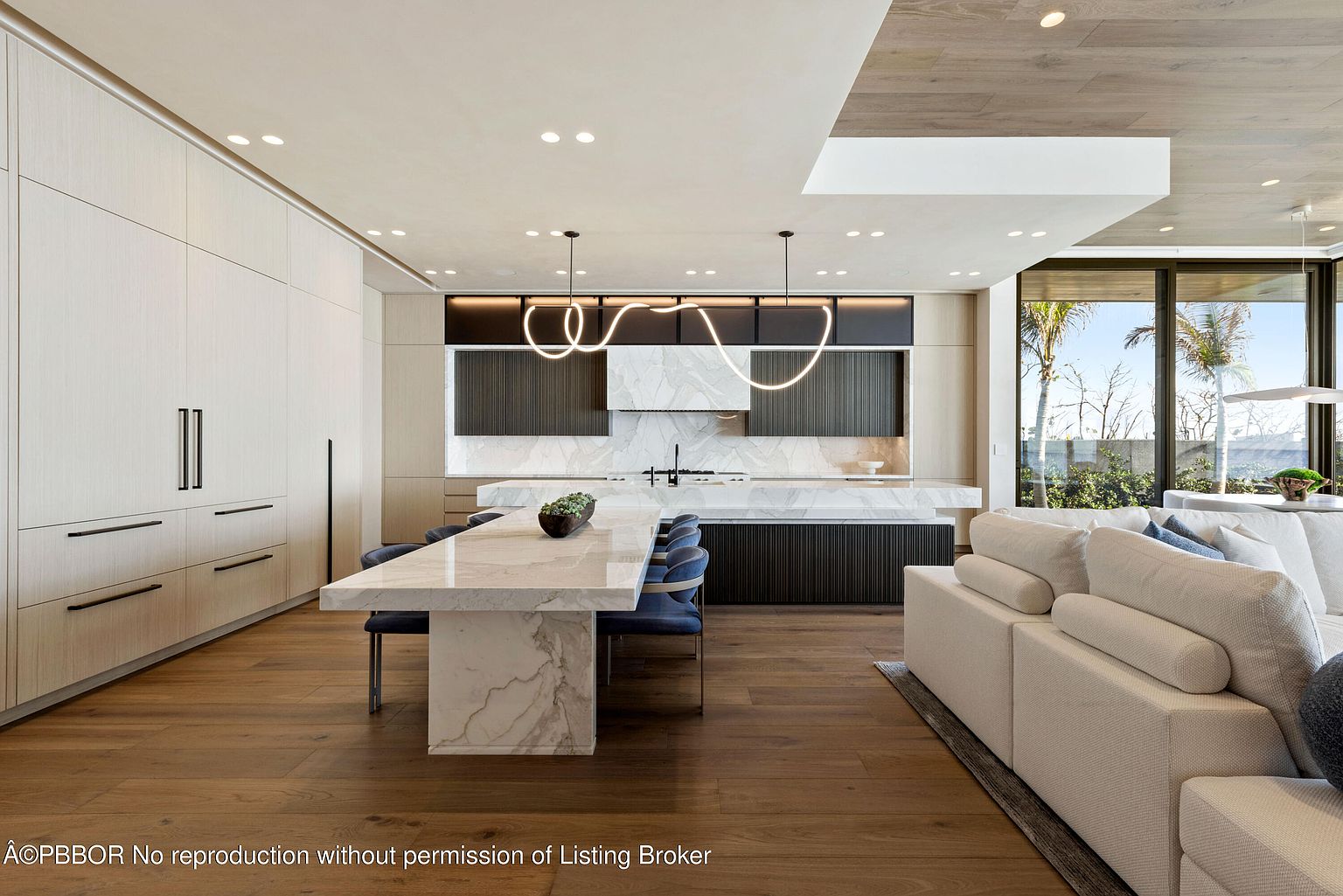 This interior shot showcases a modern kitchen and dining area with a large marble island that doubles as a dining table, surrounded by blue chairs. The kitchen features sleek, minimalist cabinetry in light wood tones and dark accents, complemented by unique, looping pendant lights. A glimpse of the living area with a comfortable white sofa is visible, creating an open and inviting space.