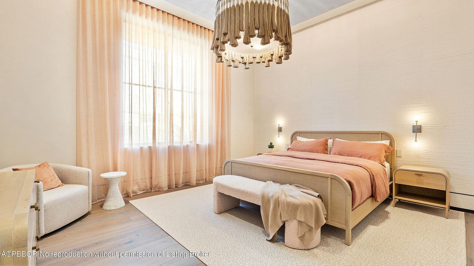 This is a bright and airy primary bedroom featuring a neutral color palette with peach accents. The room is well-lit by natural light filtering through sheer curtains, complemented by a unique chandelier and wall sconces. A woven bed frame and bench add texture, while the textured walls provide a subtle, sophisticated backdrop.