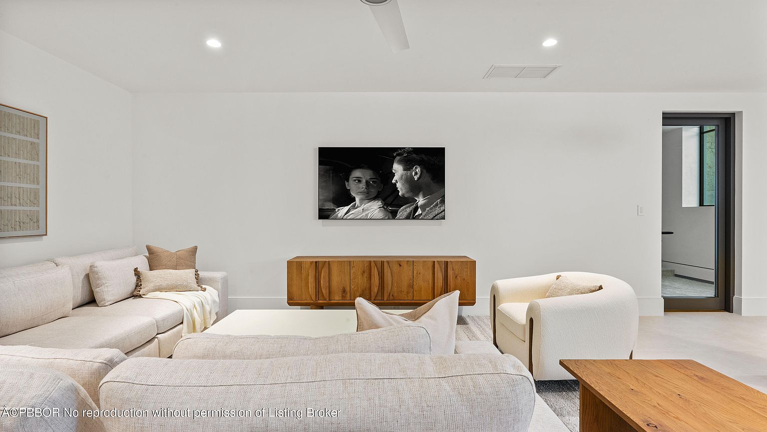 This is a bright and airy living room featuring a neutral color palette with white walls and light-colored furniture. The room is furnished with a sectional sofa, an armchair, and wooden coffee tables. A black and white photograph hangs above a wooden console, adding a touch of sophistication to the space.