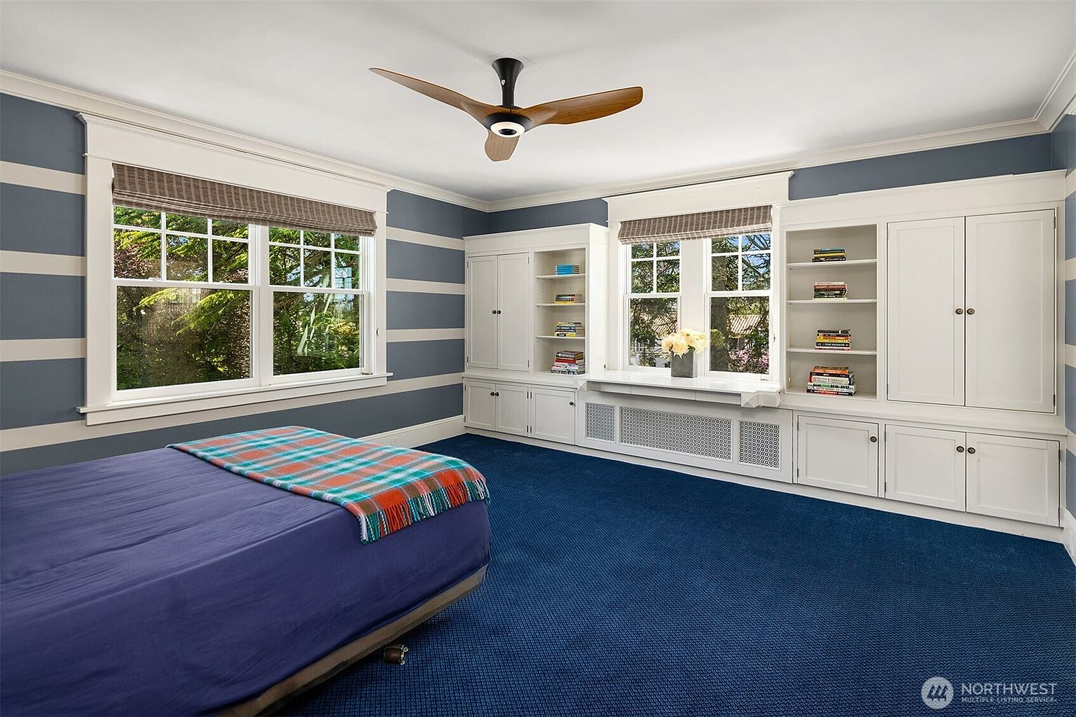 This bedroom features bold horizontal blue and white striped walls and deep blue carpeting, creating a distinct, nautical-inspired aesthetic. The room is equipped with extensive built-in white cabinetry and shelving that frames the windows, providing ample storage and display space. A modern ceiling fan with wood-toned blades centers the room, which is furnished with a bed dressed in a blue and plaid patterned comforter.