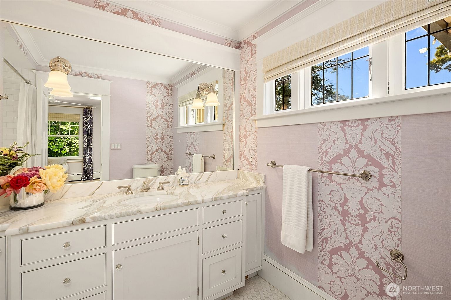 This bright bathroom features a vanity with a stone countertop and white cabinetry, complemented by a large wall-to-wall mirror. The walls are adorned with a distinctive pink and white damask-style wallpaper, creating a classic and elegant aesthetic. A window with a woven shade allows natural light to enter, while a towel rack holds a crisp white towel near the vanity.