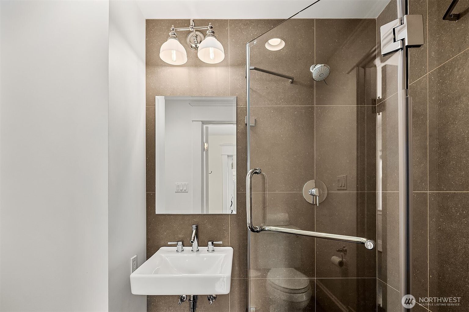 This compact, modern bathroom features a wall-mounted white sink and a glass-enclosed shower stall with a rainfall showerhead. The walls are finished with large, neutral-toned tiles that extend throughout the shower area, creating a cohesive and contemporary aesthetic. A simple vanity mirror and a two-light fixture above it provide clean, functional illumination for the space.
