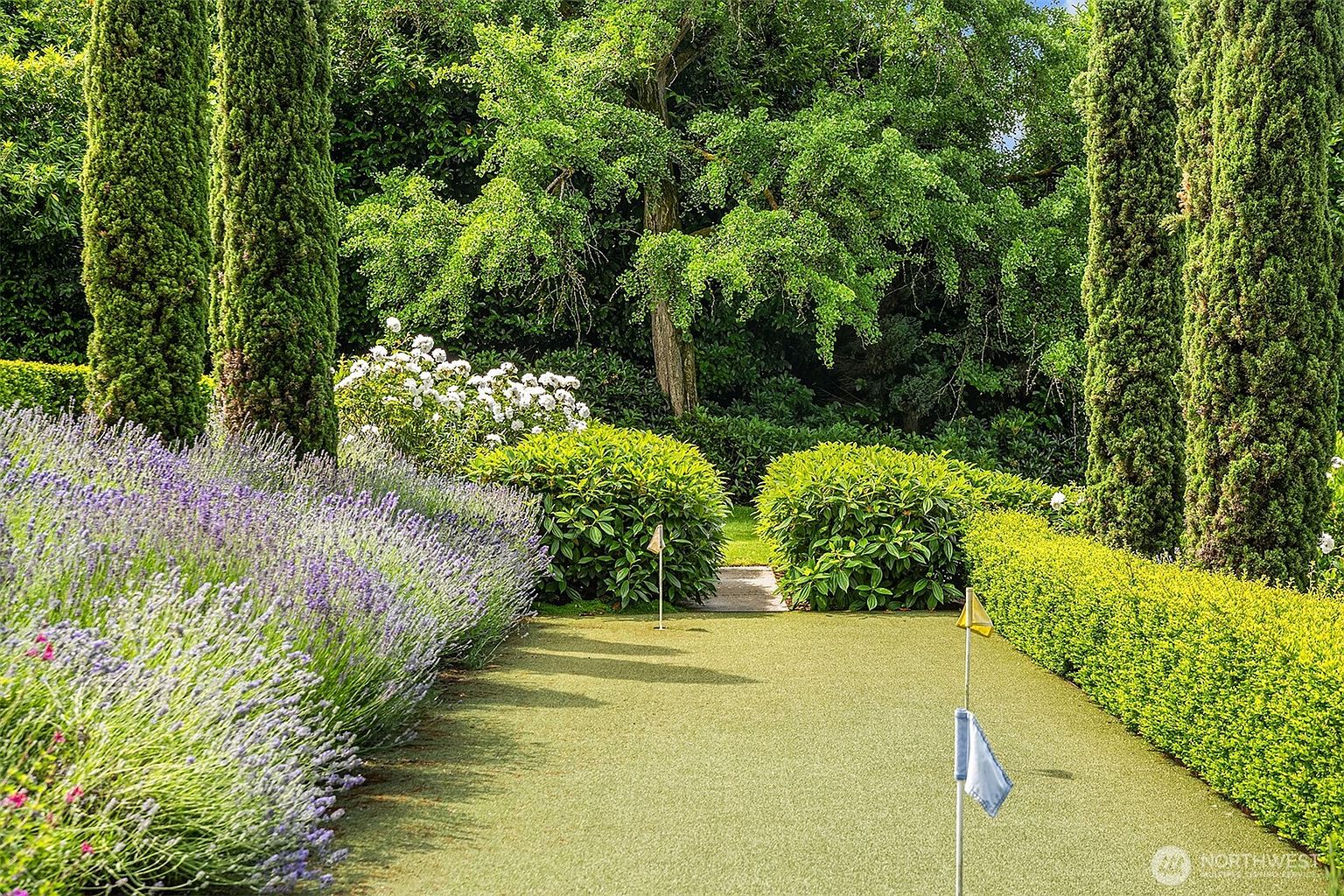 This beautifully landscaped backyard features a private putting green bordered by lush lavender bushes and manicured hedges. Tall, slender cypress trees frame the scene, leading the eye toward a small stone path nestled between dense green shrubs. The overall impression is one of serene, high-end outdoor living with a focus on recreational elegance.