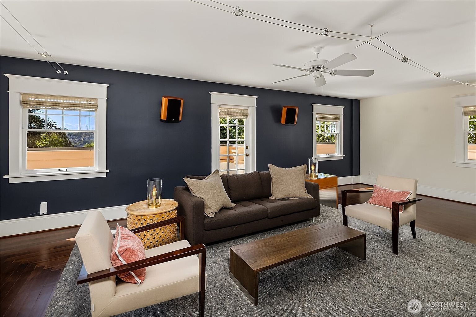 This living room features a dark navy accent wall contrasted by white trim and light-colored walls, creating a sophisticated and modern atmosphere. The space is furnished with a dark brown sofa, two cream-colored armchairs with wooden frames, and a low-profile wooden coffee table atop a textured gray area rug. A ceiling fan and track lighting provide illumination, while multiple windows and a glass-paned door allow for natural light and views of the outdoors.