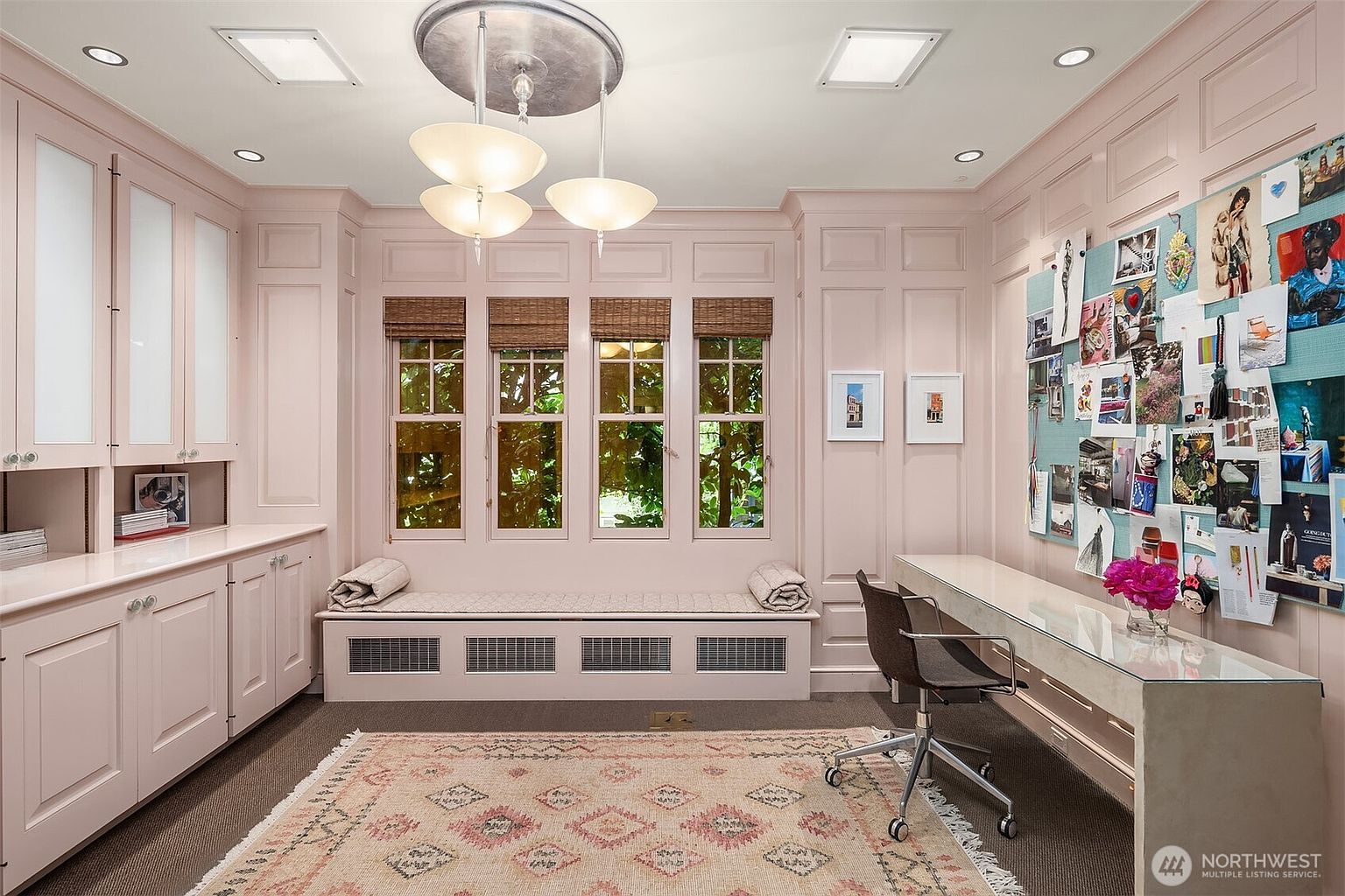 This elegant home office features soft pink paneled walls, built-in cabinetry, and a window seat nestled between four windows with woven shades. A glass-topped desk sits opposite a large pinboard wall, while a patterned area rug anchors the space under a central pendant light fixture. The room offers a sophisticated and organized workspace with a bright, inviting atmosphere.