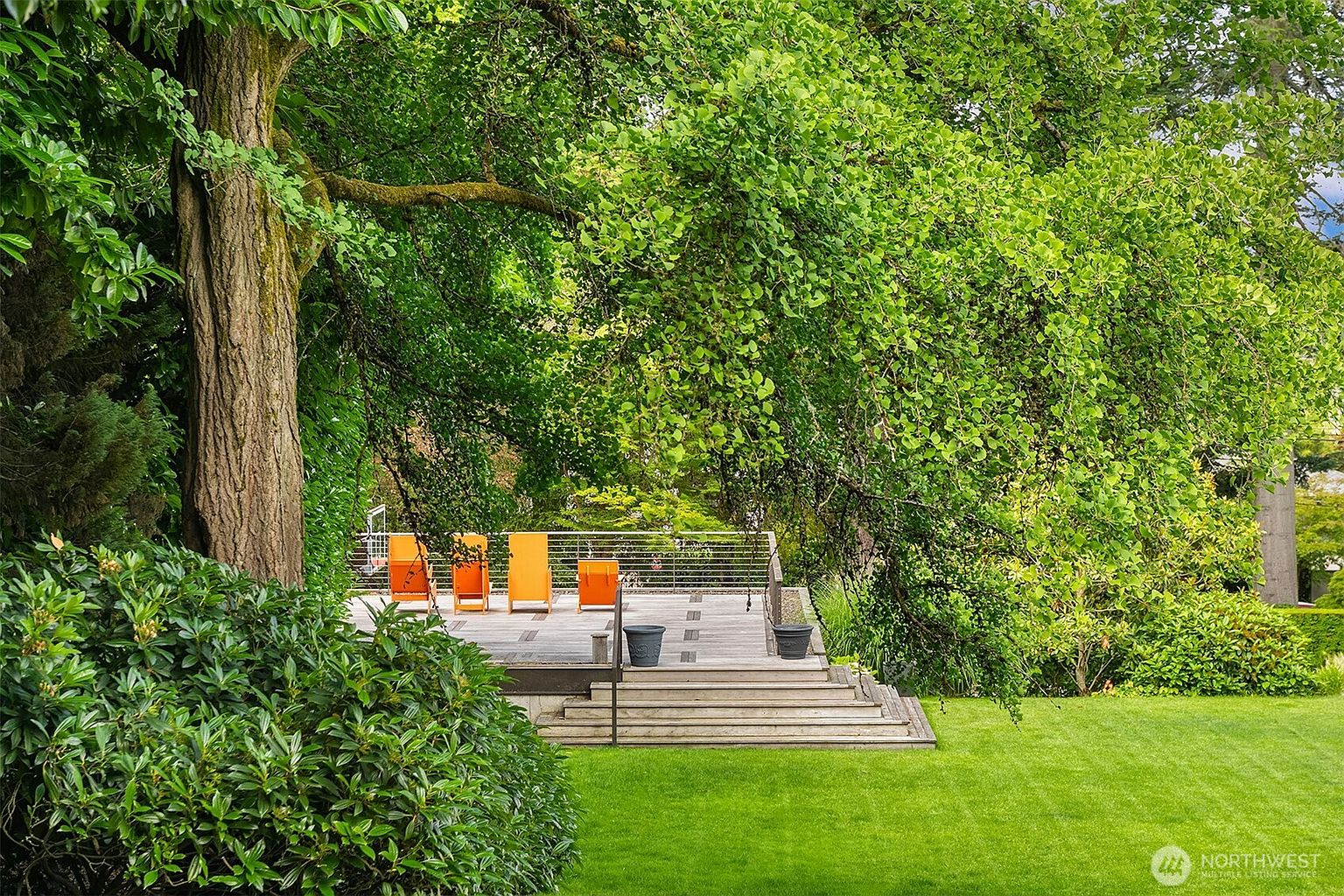 This image captures a serene outdoor deck area nestled under the lush canopy of a large, mature tree. The wooden deck features a modern cable railing and is furnished with four vibrant orange chairs, creating a bright focal point against the natural greenery. Wide, tiered wooden steps lead down to a well-manicured lawn, offering a peaceful and inviting space for relaxation.