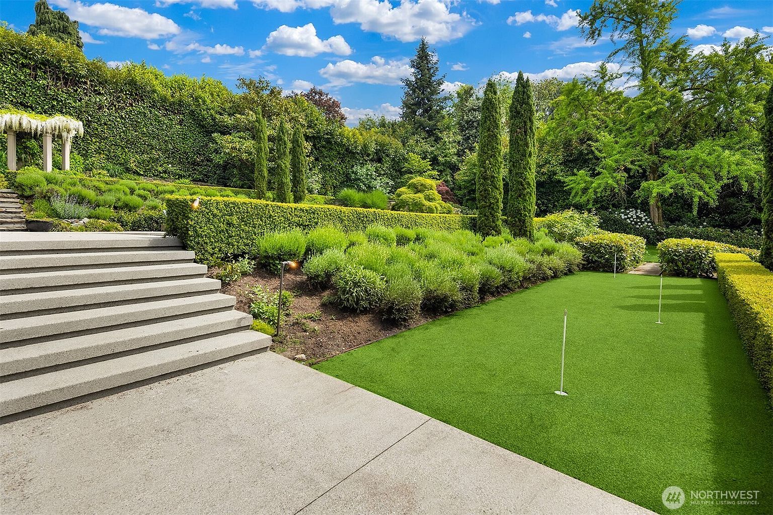 This meticulously landscaped backyard features a putting green with multiple flag pins, bordered by manicured hedges and tall, slender cypress trees. A wide set of concrete stairs leads up from a paved patio area, creating a tiered effect that emphasizes the garden's depth and structured design. The lush greenery and vibrant blue sky with scattered clouds provide a serene and upscale outdoor living environment.