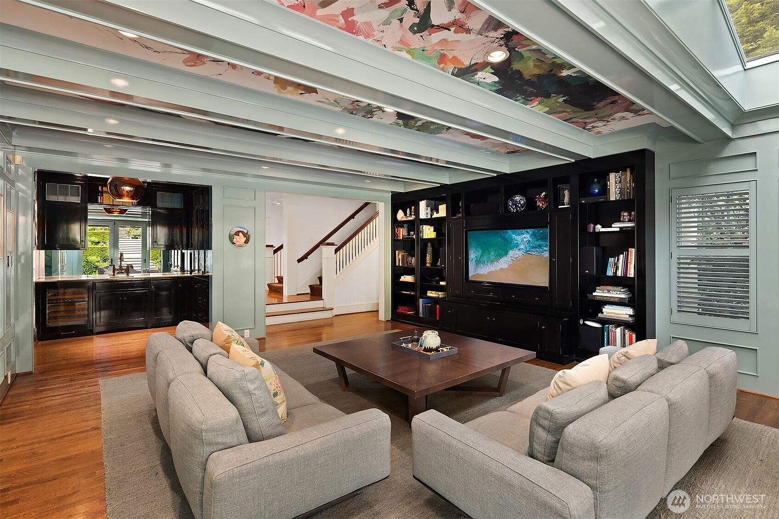 This sophisticated living room features a striking coffered ceiling with vibrant, abstract-patterned wallpaper panels between the beams. A large, dark-toned built-in media center and bookshelf unit anchors the room, while two plush gray sofas face a central wooden coffee table. The space flows seamlessly into an adjacent wet bar area and a staircase, creating an open and inviting atmosphere with a blend of modern and classic design elements.