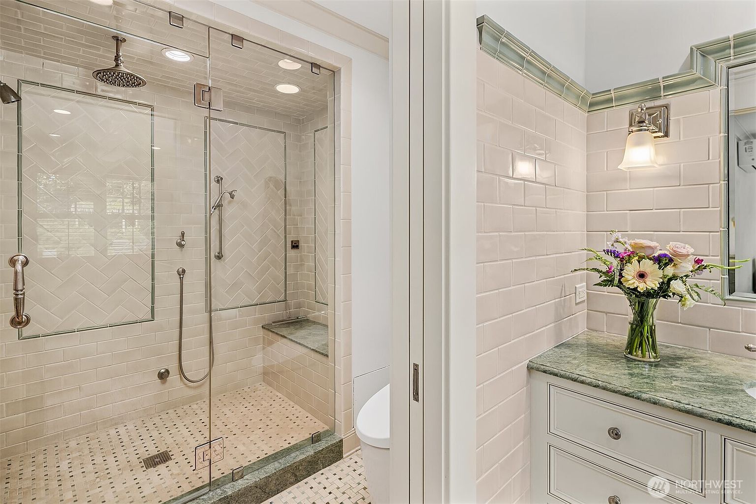 This elegant bathroom features a glass-enclosed walk-in shower with herringbone-patterned wall tiles and a built-in stone bench. Adjacent to the shower, a vanity with a green stone countertop and a decorative floral arrangement adds a touch of sophistication. The space is finished with classic subway tiles on the walls and a decorative border, creating a bright and polished aesthetic.