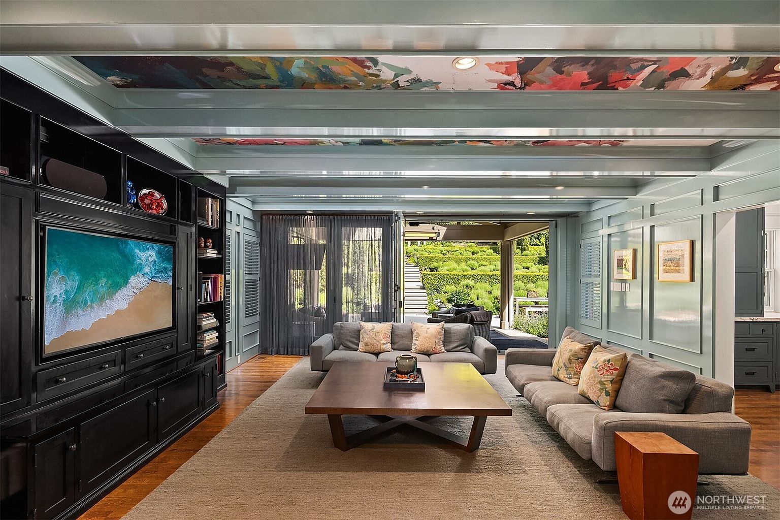 This sophisticated living room features a striking ceiling with a vibrant, multi-colored abstract mural set between light blue beams. The space is anchored by a large, dark built-in media center and two plush grey sofas arranged around a substantial wooden coffee table. Large glass doors open to a lush, terraced garden, creating a seamless transition between the elegant interior and the outdoor landscape.