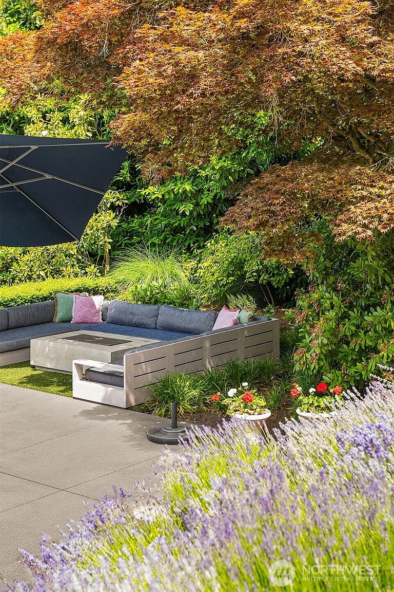 This inviting outdoor living space features a modern, low-profile sectional sofa arranged around a square concrete fire pit on a paved patio. The area is shaded by a large dark umbrella and surrounded by lush, mature landscaping, including a vibrant Japanese maple and blooming lavender in the foreground. The scene offers a serene and private atmosphere, perfect for relaxation or entertaining in a well-maintained garden setting.