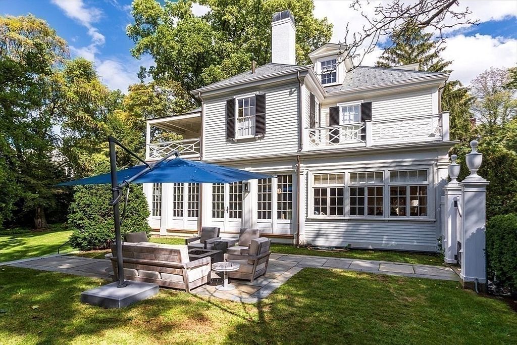 This image captures the rear exterior of a classic, light-gray clapboard home, showcasing a well-manicured lawn and a stone patio furnished with comfortable outdoor seating. A large blue cantilever umbrella provides shade over the lounge area, while the house features elegant architectural details like white-trimmed windows, a second-story balcony, and a prominent chimney. The perspective is at ground level, emphasizing the inviting transition between the indoor living space and the lush, tree-lined backyard.