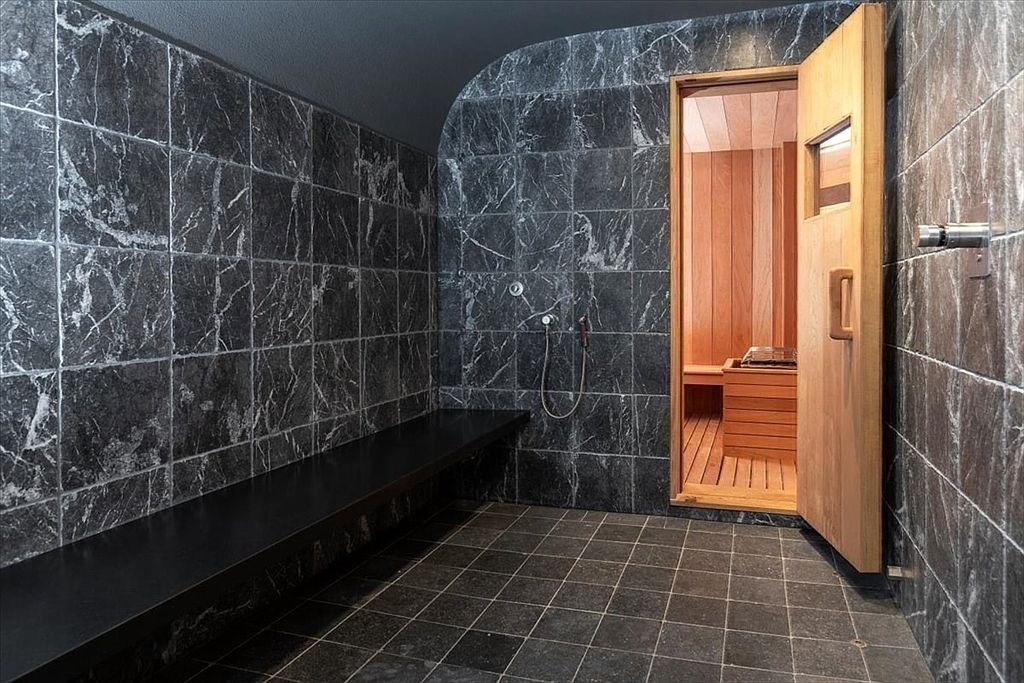 This luxurious spa-inspired bathroom features dark, veined marble wall tiles and a sleek, minimalist black bench seat. A wooden sauna door stands slightly ajar, revealing a warm, cedar-lined interior with a traditional heater, creating a sophisticated contrast between the cool stone and warm wood. The perspective is eye-level, emphasizing the high-end materials and the seamless transition between the shower area and the private sauna.