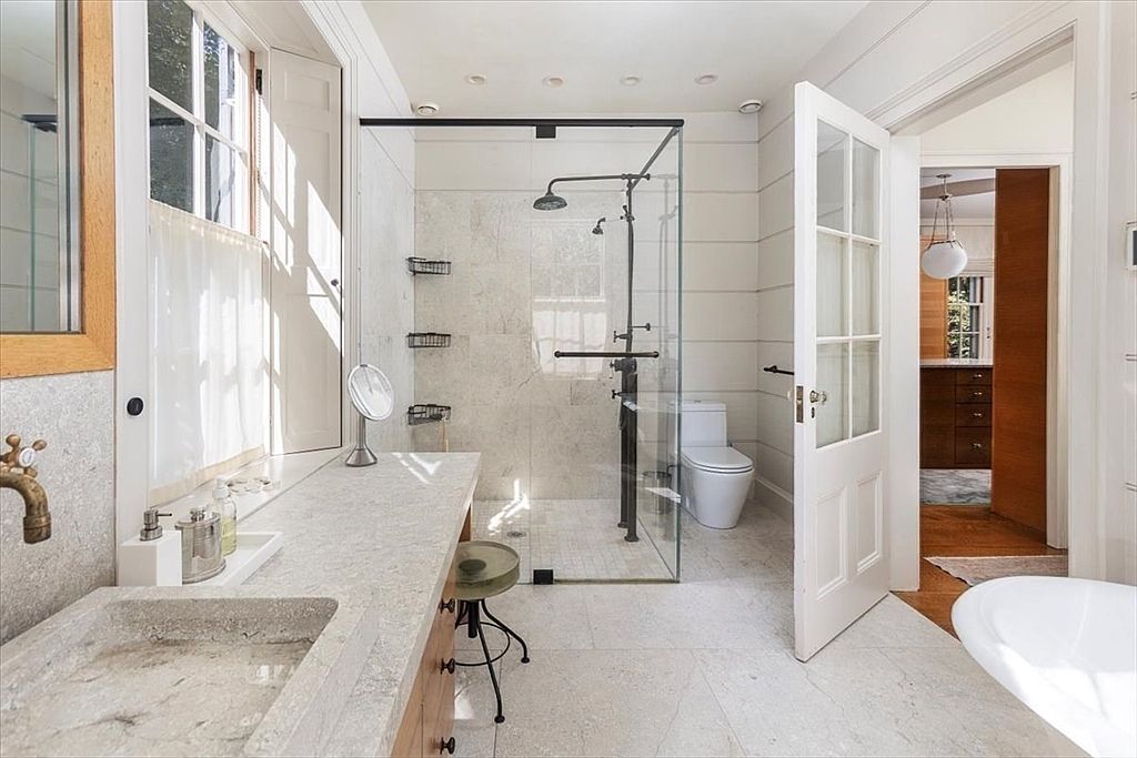 This elegant primary bathroom features a stone-topped vanity with a rustic stone sink and a glass-enclosed walk-in shower with a rainfall showerhead. The space is brightened by natural light from a large window and includes a modern toilet, all set against a backdrop of neutral, textured walls and light stone flooring. The perspective is a wide-angle shot from the doorway, showcasing the seamless blend of rustic materials and contemporary design.