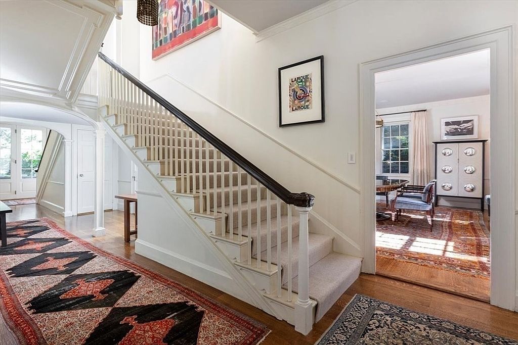 This elegant foyer features a grand staircase with a dark wood handrail and white balusters, set against a backdrop of light-filled, neutral walls. A large, vibrant abstract painting hangs above the stairs, while intricate patterned rugs define the hardwood floor space. The perspective looks from the entryway toward a dining room visible through an open doorway, creating a sense of depth and sophisticated flow.