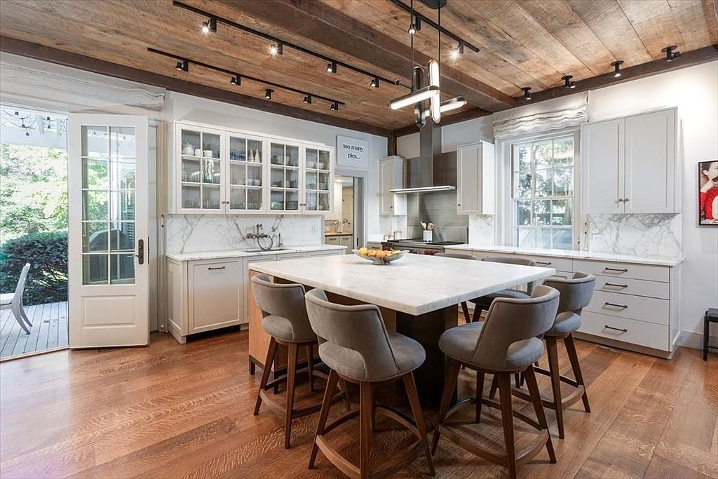 This elegant, rustic-modern kitchen features a large central island with a marble countertop and four upholstered bar stools, serving as the focal point of the room. The space is defined by a striking reclaimed wood ceiling with exposed beams and track lighting, complemented by white cabinetry and marble backsplashes. A glass-paned door opens to an outdoor deck, creating a seamless transition between the bright, airy interior and the natural surroundings.