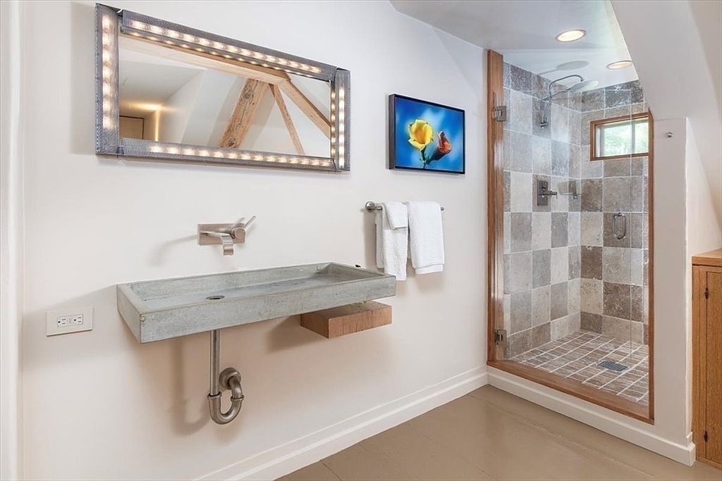 This modern bathroom features a unique, industrial-style concrete wall-mounted sink paired with a sleek, illuminated mirror. To the right, a walk-in shower is enclosed by a glass door and finished with textured, multi-toned stone tiles. The overall aesthetic blends rustic, raw materials with contemporary design elements, creating a sophisticated and minimalist atmosphere.