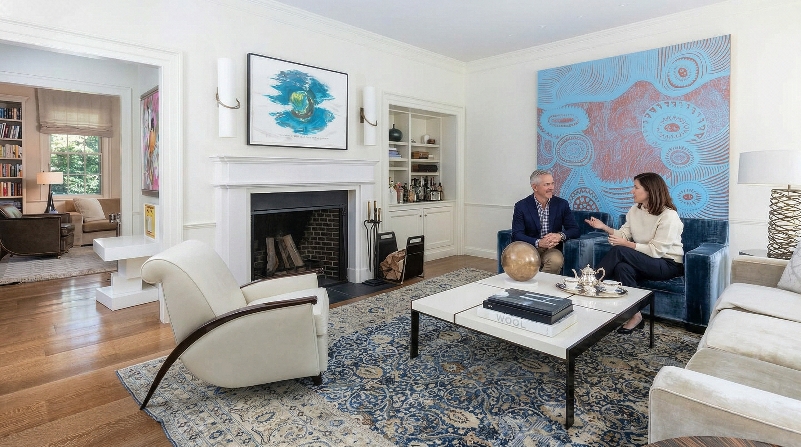 This sophisticated living room features a blend of classic and contemporary design, anchored by a large, ornate blue-toned area rug and a striking abstract mural on the wall. The space includes a white fireplace mantel, plush blue velvet armchairs, and a sleek white coffee table, all set against a backdrop of hardwood floors and elegant built-in shelving. The perspective is a wide-angle shot from the room's entrance, showcasing the open flow into an adjacent library area.