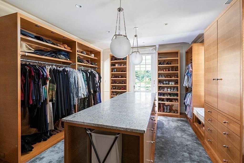 This spacious, high-end walk-in closet features custom light-wood cabinetry, extensive hanging space, and dedicated shelving for shoes and accessories. A large central island with a granite countertop provides a functional workspace, while elegant globe pendant lights hang from the ceiling, creating a sophisticated and organized atmosphere. The room is finished with a plush grey rug and leads to a bright glass-paned door, offering a view of the outdoors.