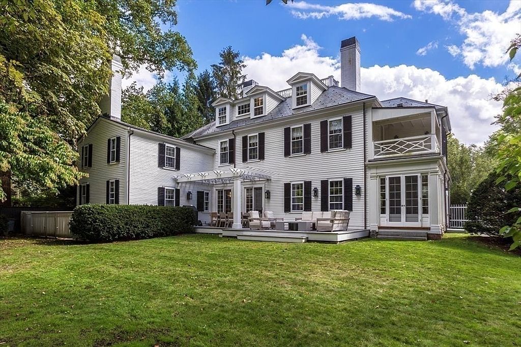 This image captures the rear exterior of a stately, white-sided colonial home featuring a classic architectural style with black shutters and multiple dormer windows. A spacious wooden deck with a pergola and comfortable outdoor seating provides an inviting space for relaxation, overlooking a well-manicured lawn surrounded by mature trees. The perspective is a wide, eye-level shot that emphasizes the home's elegant proportions and its seamless integration with the lush, private backyard.