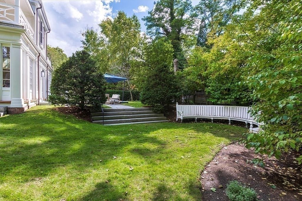 This serene backyard scene features a lush green lawn, a curved white wooden bench, and stone steps leading to a patio area with an umbrella. The perspective is from the lawn looking toward the side of a classic white home, framed by mature trees and foliage. The overall impression is one of a peaceful, well-manicured private retreat perfect for outdoor relaxation.