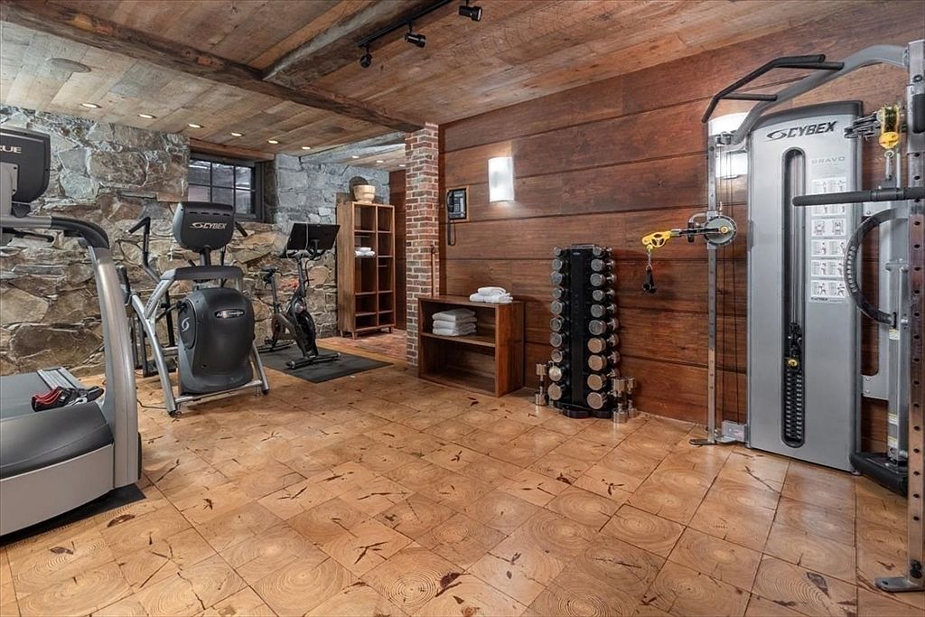 This rustic-inspired home gym features a unique floor made of wood cross-sections, complemented by stone walls and warm wood-paneled accents. The space is well-equipped with professional-grade Cybex fitness machines, including a treadmill, elliptical, and a functional trainer, alongside a rack of dumbbells. The combination of natural textures and high-end equipment creates an inviting and sophisticated workout environment.
