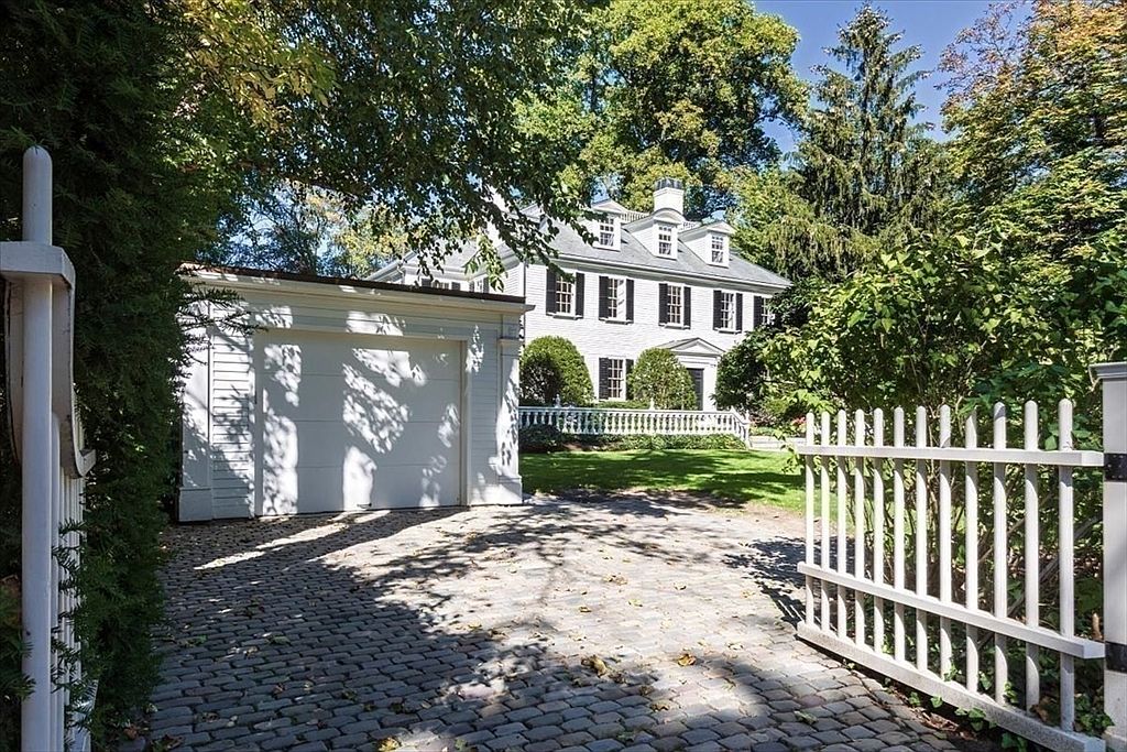 This image captures a charming, traditional-style white home viewed through an open white picket gate. The property features a cobblestone driveway leading to an attached garage, with a lush green lawn and mature trees framing the elegant, multi-story residence. The perspective is from the entrance, inviting the viewer into the serene and well-manicured grounds of the estate.