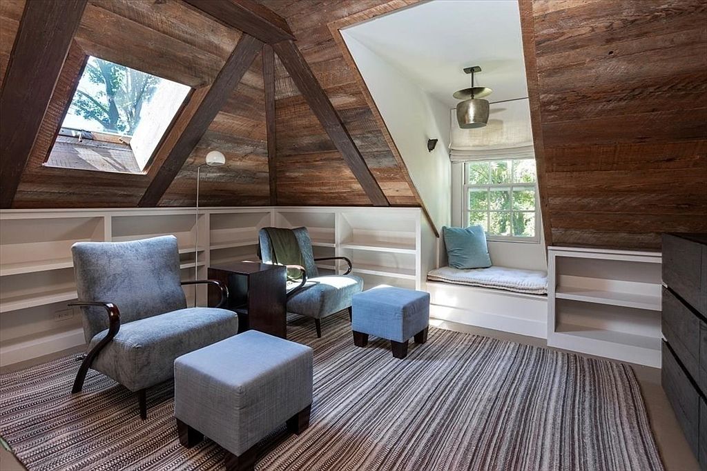 This cozy attic space features rustic wood-paneled walls and exposed ceiling beams, creating a warm and inviting atmosphere. The room is furnished with two comfortable blue armchairs, matching ottomans, and a built-in window seat, all arranged on a striped area rug. Built-in white shelving lines the walls, maximizing storage in the sloped space, while a skylight and a window provide ample natural light.