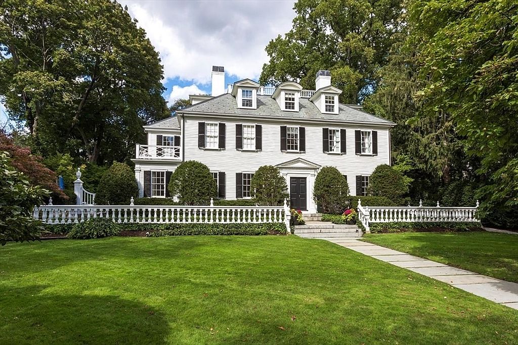 This stately, white colonial-style home features a symmetrical facade with dark shutters, a central front door, and three dormer windows on the roof. A manicured lawn leads up to a decorative white balustrade fence, while mature trees frame the property, creating a sense of privacy and elegance. The perspective is a straight-on, eye-level shot that captures the grand scale and classic architectural charm of the residence.
