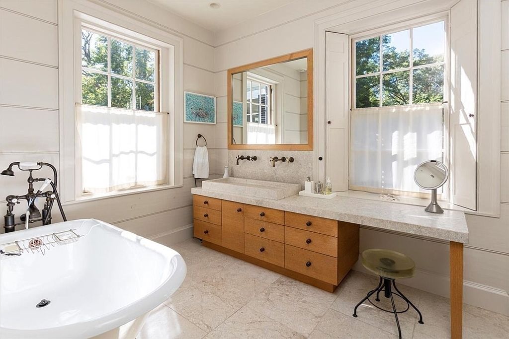 This elegant primary bathroom features a light-filled, airy aesthetic with a custom stone vanity, warm wood cabinetry, and a freestanding soaking tub. The space is anchored by large windows with classic shutters, creating a serene and spa-like atmosphere. The neutral color palette and clean lines offer a sophisticated, timeless design perfect for a luxury home.
