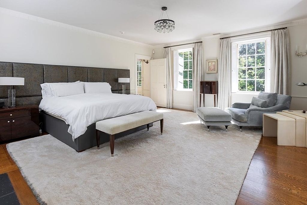 This spacious primary bedroom features a large bed with a dark, tufted headboard, complemented by a plush, light-colored area rug covering the hardwood floors. The room is brightened by two large windows with elegant floor-to-ceiling curtains, a cozy armchair with an ottoman, and a sophisticated crystal chandelier. The overall aesthetic is one of refined luxury and calm, offering a serene retreat with ample natural light.