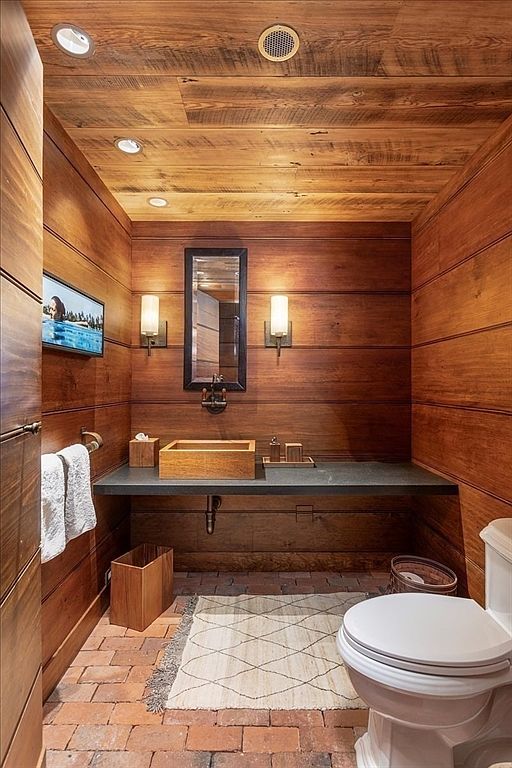 This rustic-style powder room features warm, horizontal wood-paneled walls and a matching wood-plank ceiling, creating a cozy, cabin-like atmosphere. The space is anchored by a dark stone countertop with a unique wooden vessel sink, complemented by a brick floor and a textured area rug. A wall-mounted television, elegant sconce lighting, and a framed mirror complete this sophisticated yet earthy bathroom design.