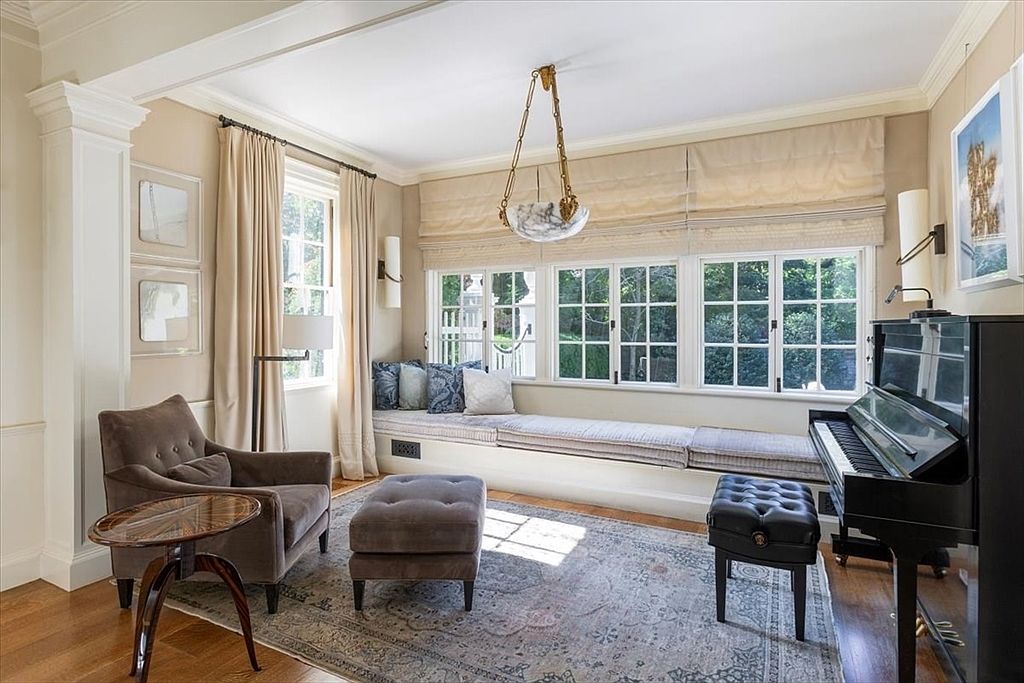 This elegant living room features a cozy window seat lined with plush cushions, a grand piano, and a comfortable armchair with an ottoman. The space is bathed in natural light from large windows adorned with soft Roman shades and floor-to-ceiling curtains, creating a sophisticated and inviting atmosphere. A decorative chandelier hangs centrally, complementing the traditional architectural details and the patterned area rug that anchors the room.