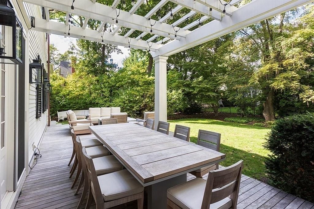 This spacious wooden deck features a large, rustic dining table surrounded by comfortable chairs, perfect for outdoor entertaining. A white pergola overhead provides partial shade and is adorned with string lights, while a separate lounge area with plush seating is visible in the background. The deck overlooks a lush, green backyard, creating a serene and inviting atmosphere for relaxation.