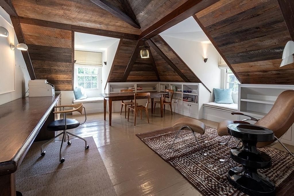 This spacious attic office features a warm, rustic aesthetic with exposed wooden beams and planked ceilings. The room is well-organized with a long built-in desk, a central dining-style table for collaborative work, and cozy reading nooks tucked into the dormer windows. A patterned area rug anchors the seating area, which includes a leather lounge chair and ottoman, creating a sophisticated yet comfortable workspace.