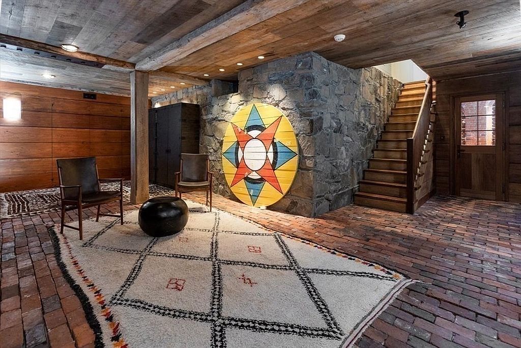 This rustic basement features a unique blend of textures, including exposed brick flooring, reclaimed wood ceilings, and a rugged stone accent wall. A large, colorful geometric art piece serves as a focal point, complemented by mid-century modern leather chairs and a patterned area rug. The space is illuminated by recessed lighting, creating a warm and inviting atmosphere that feels both historic and curated.
