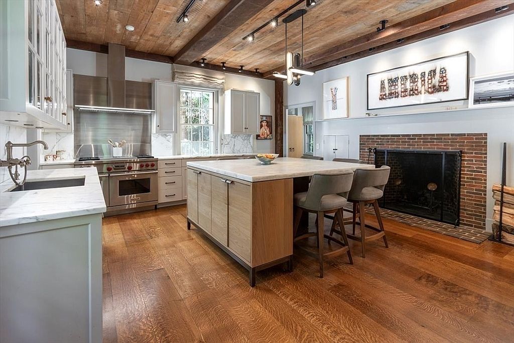 This sophisticated kitchen blends rustic charm with modern luxury, featuring a warm wood-plank ceiling with exposed beams and rich hardwood flooring. A large central island with a marble countertop serves as the focal point, complemented by stainless steel appliances, white cabinetry, and a cozy brick fireplace. The perspective is a wide-angle shot that captures the inviting, open-concept layout, highlighting the seamless integration of cooking and living spaces.