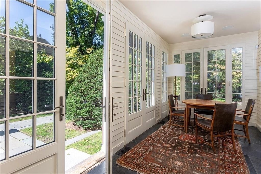 This bright and airy sunroom features floor-to-ceiling glass-paned doors that open to a lush garden, creating a seamless transition between indoor and outdoor living. The space is furnished with a classic wooden dining set atop a traditional patterned area rug, illuminated by a modern pendant light. The white shiplap walls and dark flooring provide a sophisticated, clean aesthetic perfect for casual dining or relaxation.