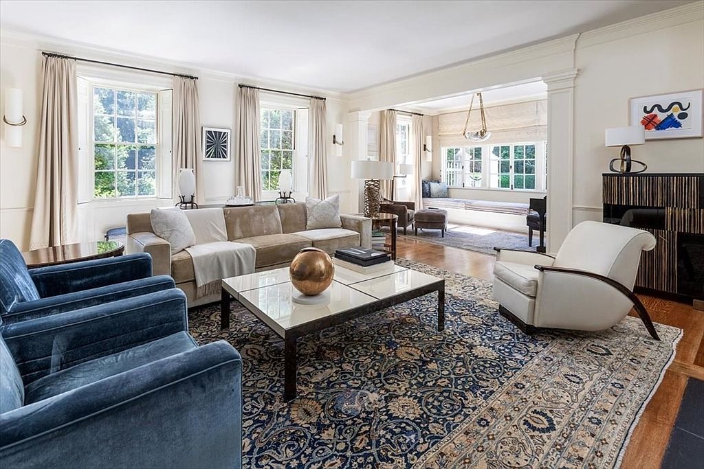 This elegant living room features a sophisticated blend of traditional and modern design, anchored by a large, ornate area rug and a central coffee table with a decorative gold sphere. The space is furnished with plush blue velvet armchairs, a neutral-toned sofa, and a sleek white accent chair, all bathed in natural light from multiple large windows. The open-concept layout flows seamlessly into a sun-drenched reading nook, creating an inviting and airy atmosphere perfect for refined entertaining.