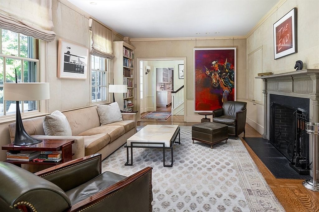 This sophisticated living room features a warm, neutral color palette with a plush beige sofa, leather armchairs, and a central marble-topped coffee table. A large, vibrant painting of a cowboy on horseback serves as a striking focal point, complemented by a traditional fireplace and a patterned area rug. The room is well-lit with natural light from large windows and offers a seamless transition into an adjacent hallway, creating an inviting and elegant atmosphere.
