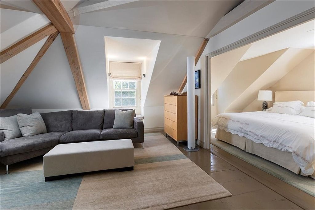 This attic space features a cozy living area with a grey sectional sofa and a matching ottoman, set beneath exposed wooden beams that highlight the vaulted ceiling. A doorway leads into an adjacent bedroom, creating a seamless flow between the relaxation and sleeping quarters. The room is bathed in natural light from a dormer window, contributing to an airy and inviting atmosphere.