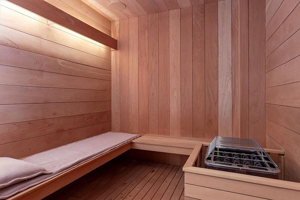 This image showcases a compact, wood-paneled sauna featuring horizontal and vertical cedar planking that creates a warm, rustic atmosphere. A built-in wooden bench with a soft cushion runs along the wall, while a metal heater unit is positioned in the corner, protected by a wooden guard rail. The perspective is from a low angle, emphasizing the cozy, intimate nature of this wellness space.