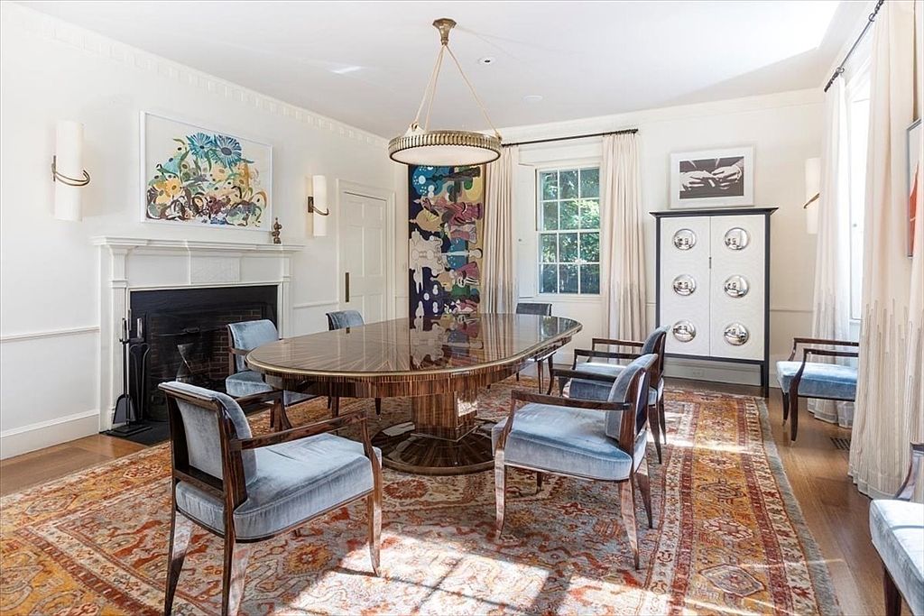 This elegant dining room features a polished, oval-shaped wooden table surrounded by plush, light blue velvet armchairs, set atop a large, intricately patterned oriental rug. The room is anchored by a grand fireplace with a vibrant abstract painting above it, while a tall, modern cabinet with circular accents stands against the wall. The space is bathed in natural light from the windows, creating a sophisticated and inviting atmosphere perfect for formal gatherings.